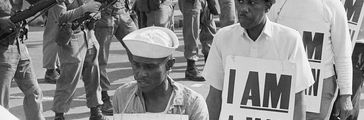National Guard troops stand with bayonets fixed as African-American sanitation workers peacefully march by while wearing placards reading "I AM A MAN." 