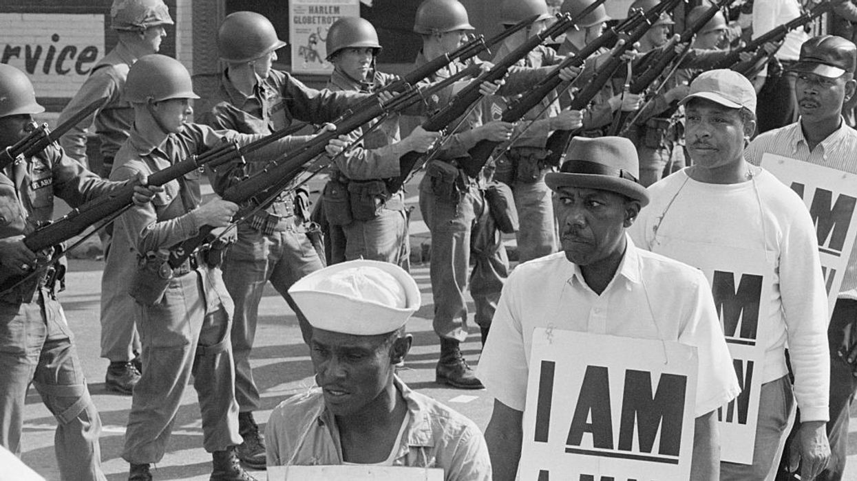 National Guard troops stand with bayonets fixed as African-American sanitation workers peacefully march by while wearing placards reading "I AM A MAN."