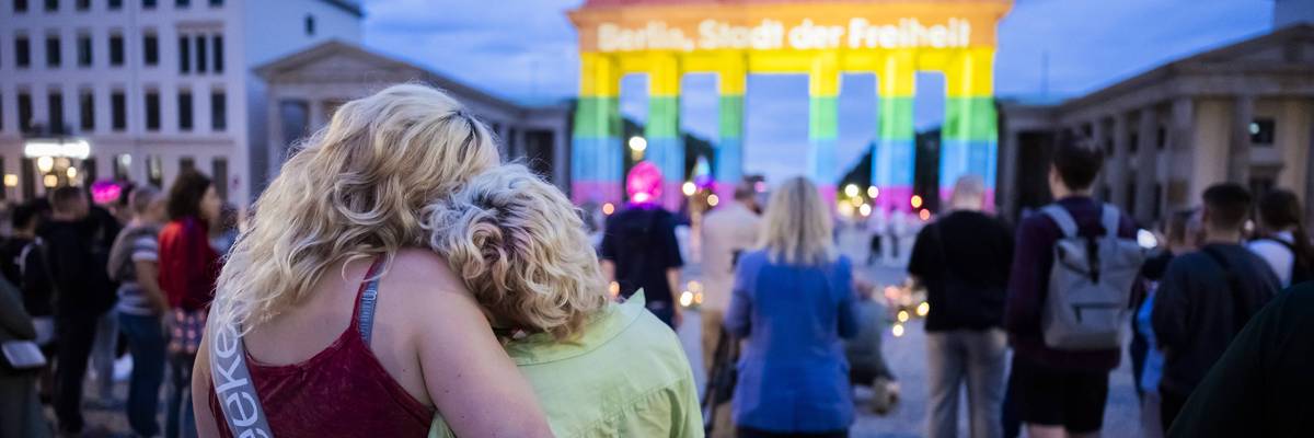 People embrace at a memorial for the Berlin Pride attack near a Brandenburg Gate illuminated in rainbow colors
