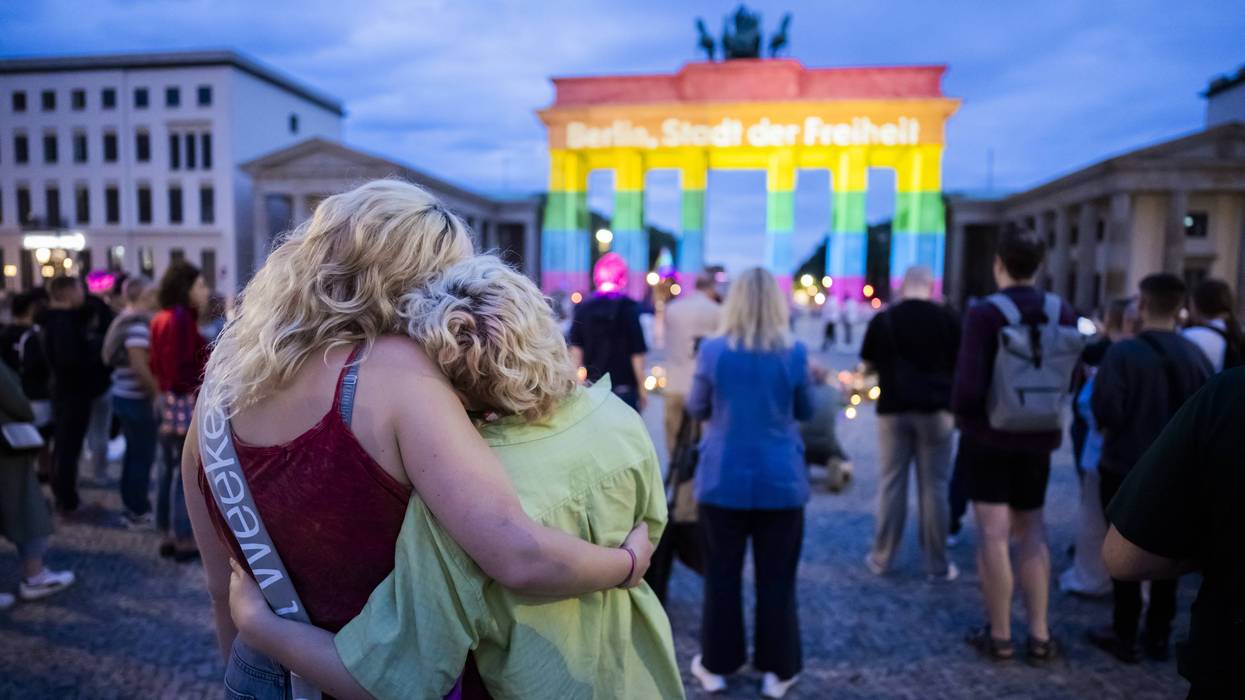 People embrace at a memorial for the Berlin Pride attack near a Brandenburg Gate illuminated in rainbow colors