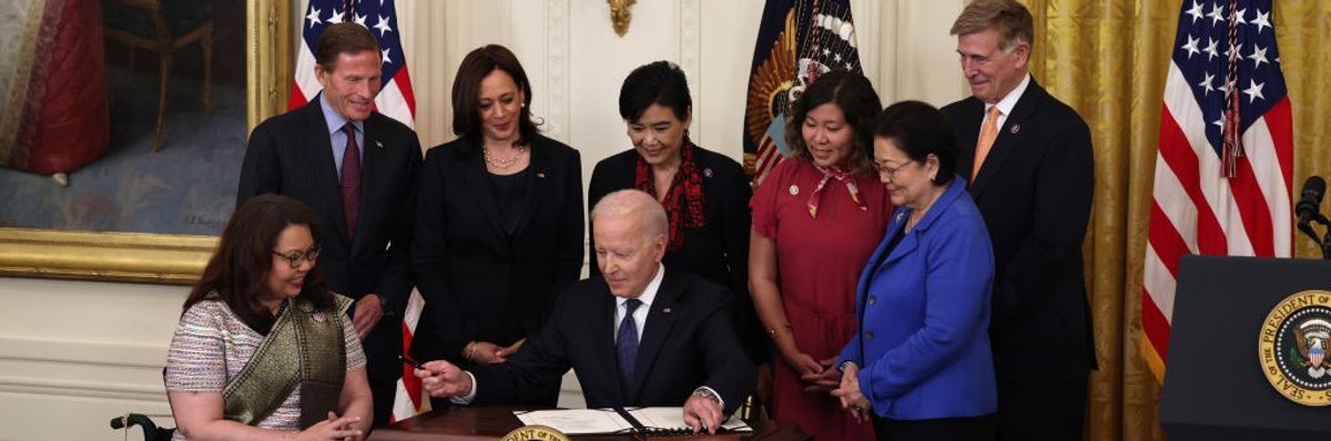 President Joe Biden sits at a desk holding a pen surrounded by members of Congress.