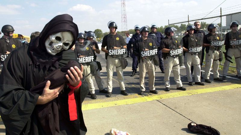 Protester dressed as death holds baby doll near Missouri Boeing plant with police behind them.
