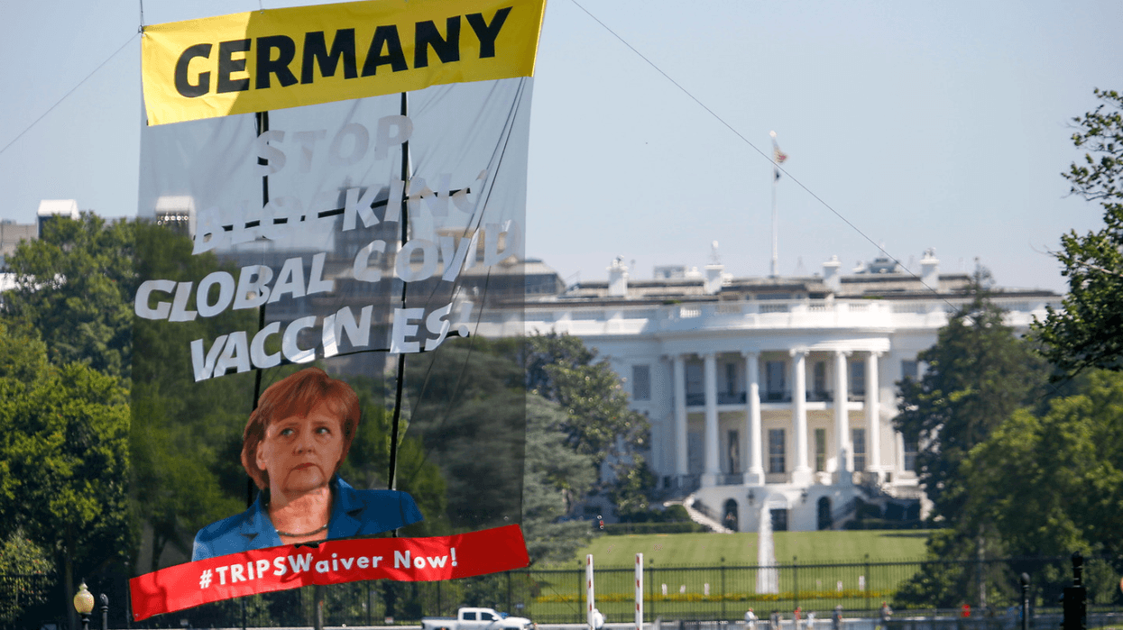 Protesters hoisted a large banner outside the White House calling on German Chancellor Angela Merkel to stop blocking an emergency waiver of intellectual property rules.