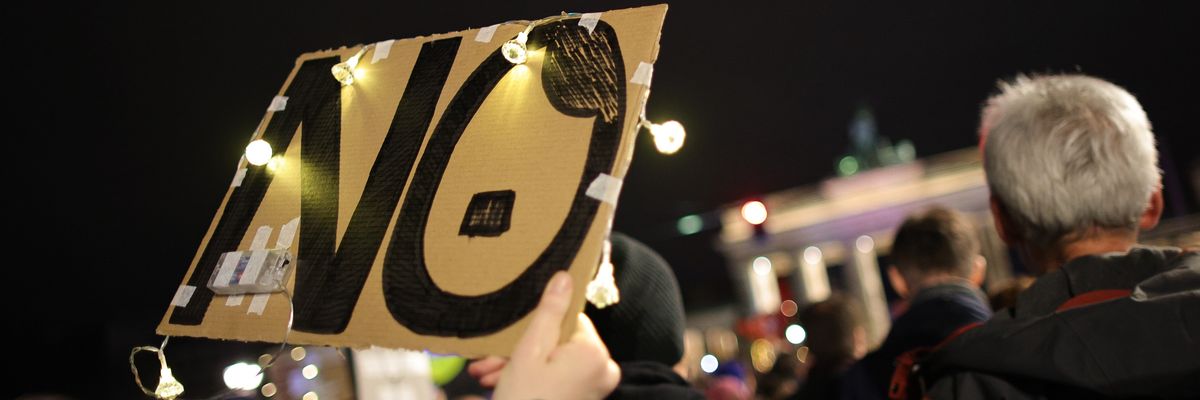 Protesters hold up a banner reading "NO" with the O shaped like an Adolf Hitler caricature
