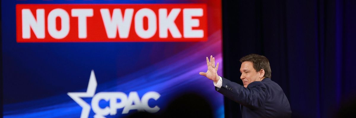 Ron DeSantis waves to a CPAC crowd by a large sign reading "not woke" 