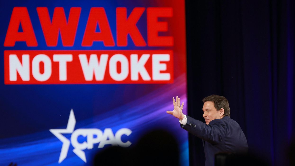 Ron DeSantis waves to a CPAC crowd by a large sign reading "not woke"