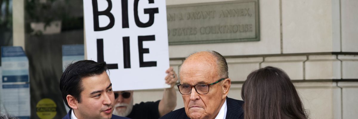 Rudy Giuliani talks to the press while a protester holds a sign reading, "Big Lie"