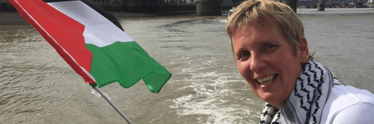 Sarah Wilkinson smiles while wearing a keffiyeh and holding a flag of Palestine.