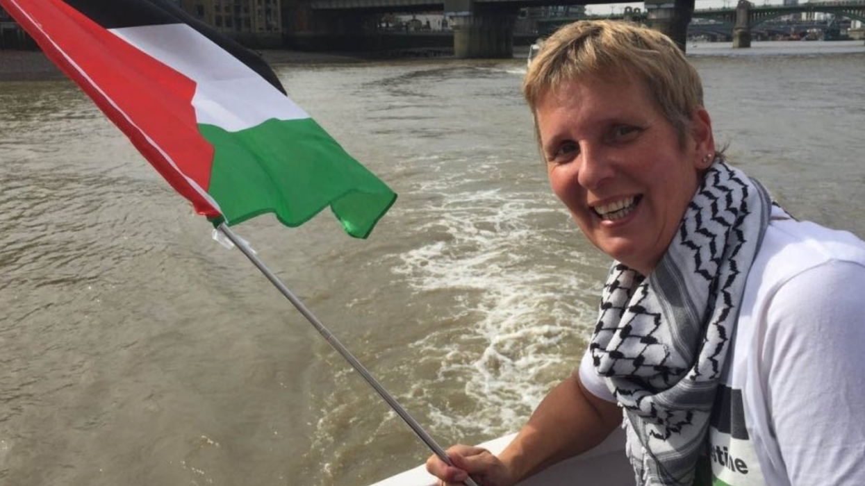 Sarah Wilkinson smiles while wearing a keffiyeh and holding a flag of Palestine.