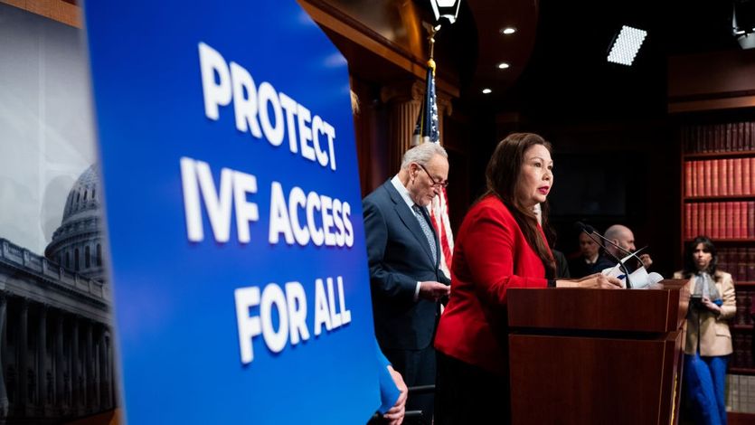 Sen. Tammy Duckworth speaks in a red suit by a sign reading "Protect IVF Access for All."