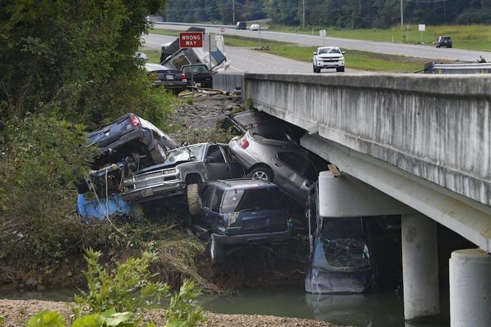 Several cars and a large pickup truck are piled up against a bridge after being swept downstream by a flood.