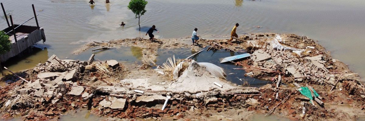 This aerial photograph taken in Pakistan's Balochistan province on August 31, 2022 shows people wading in floodwaters next to a house that collapsed due to monsoon rains turbocharged by the climate crisis.