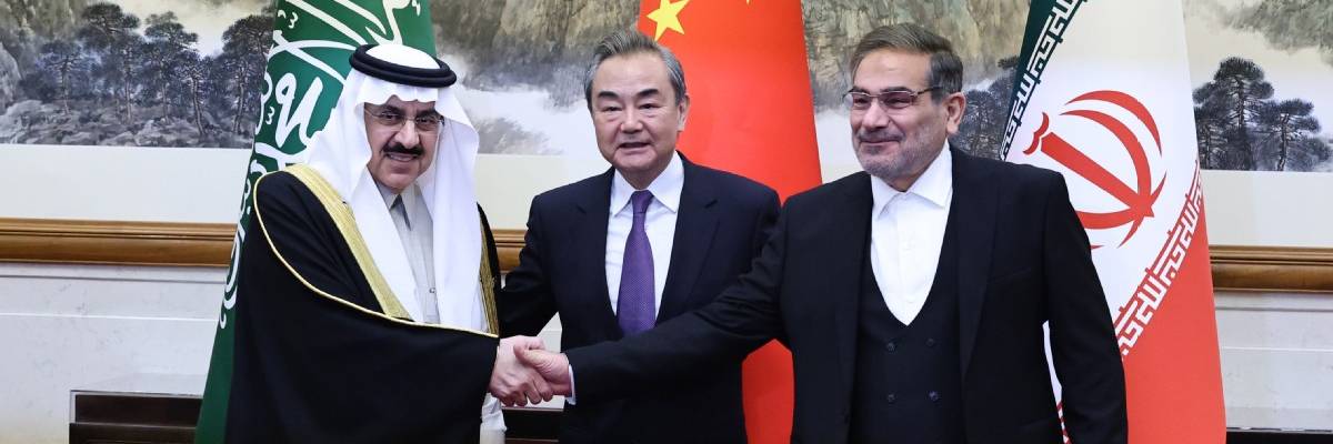 Three men shake hands in front of flags.