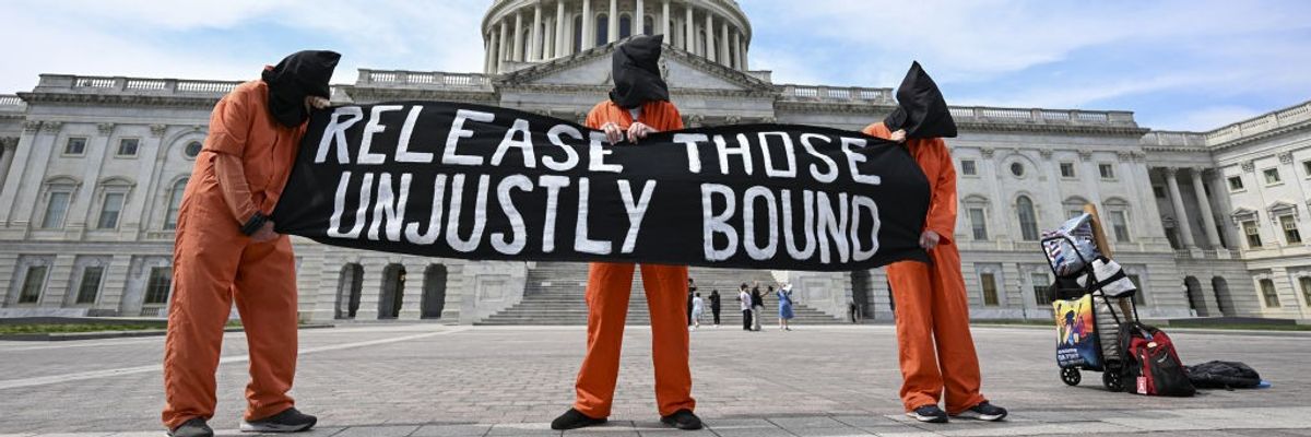 Three protesters in orange jumpsuits and black bags on their heads hold a sign reading, "Release those unjustly bound."