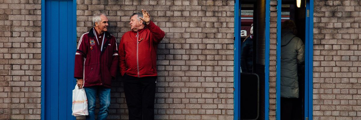 To older men lean against a brick wall talking.