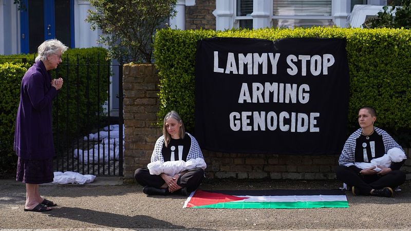 Two activists sit with models of dead babies on either side of words reading, "Lammy Stop Arming Genocide."