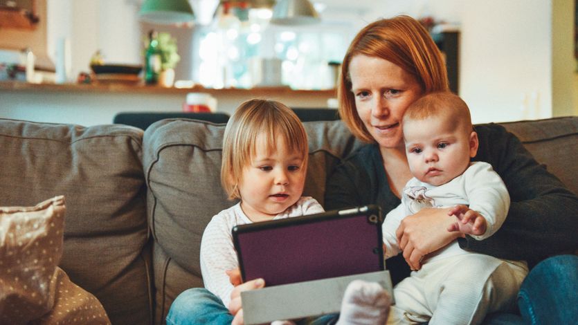 Two babies and a woman sit on a sofa while looking at a tablet. 