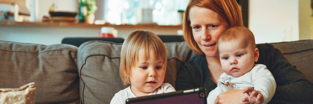 Two babies and a woman sit on a sofa while looking at a tablet.