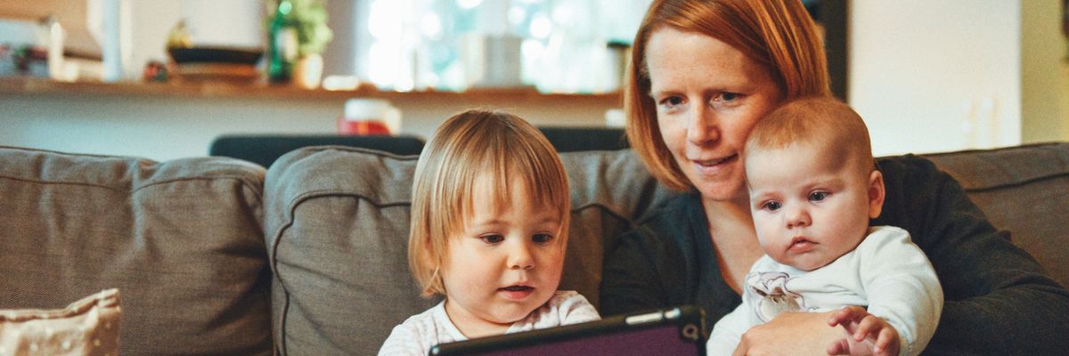 Two babies and woman sitting on sofa while holding baby and watching on tablet.