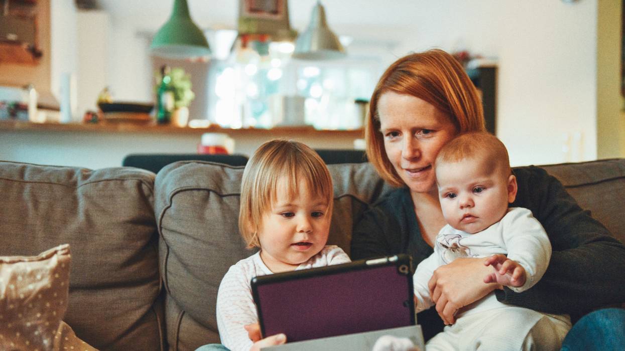 Two babies and woman sitting on sofa while holding baby and watching on tablet.