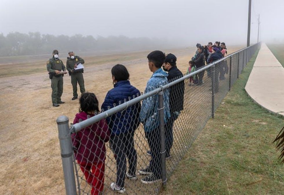 Two border patrol agents holding papers look at people standing on a dirt road in two separate groups along a fence line