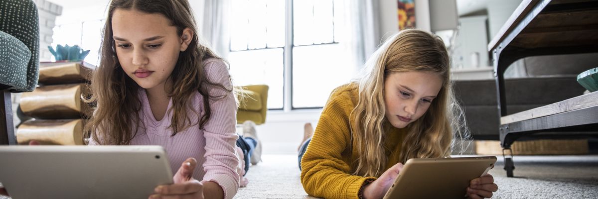 Two girls use digital tablets on a living room floor.