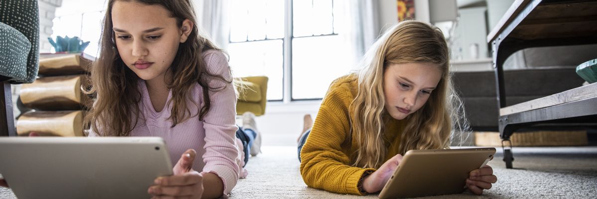 Two girls use digital tablets on a living room floor.