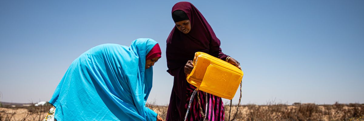 Two woman pour water from a yellow plastic tub.