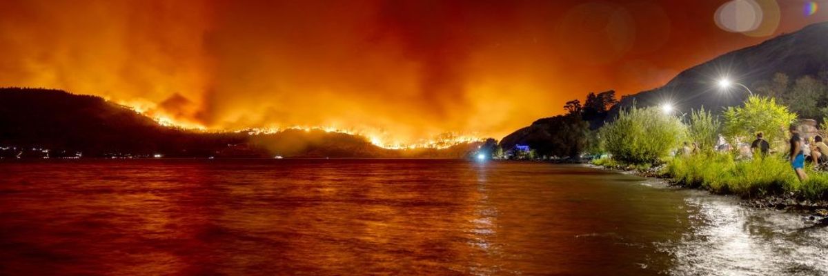 Wildfires rage above a hillside and are reflected in water.