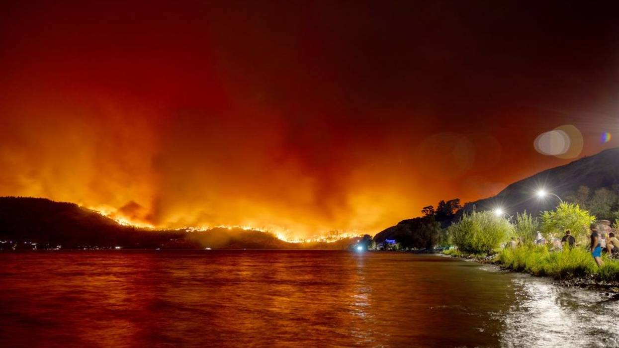 Wildfires rage above a hillside and are reflected in water.