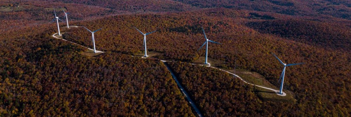 Windmills rise out of forested mountains. 