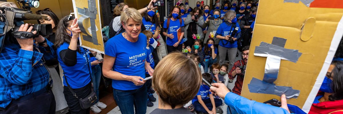 Women and children in blue shirts speak to a woman in a gray suit.