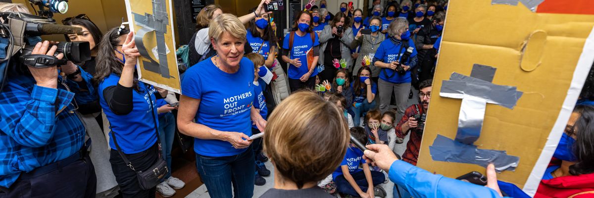 Women and children in blue shirts speak to a woman in a gray suit. 