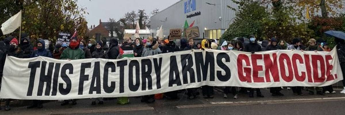 Workers protest a factory with a sign reading, "This factory arms genocide."