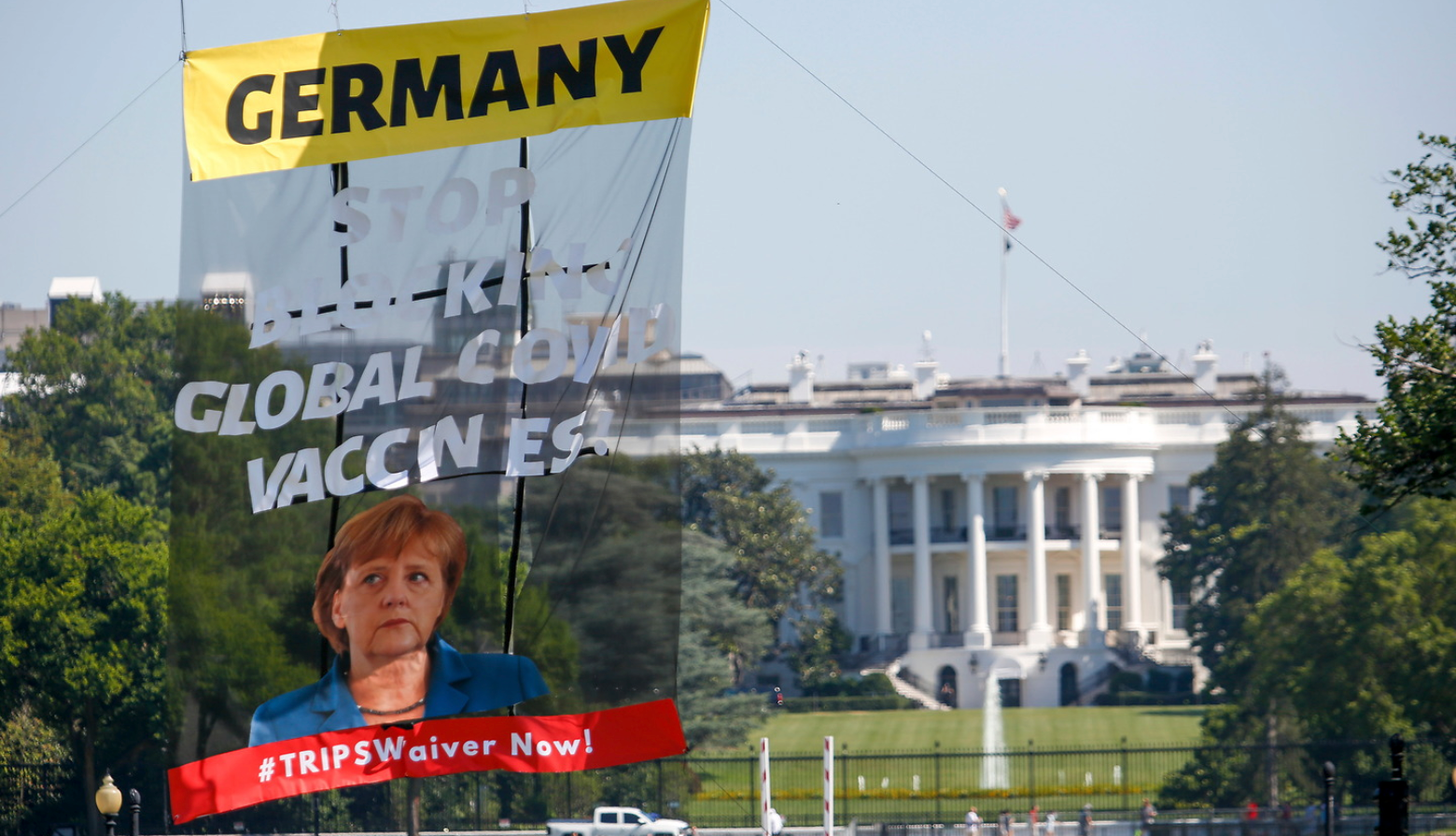 Protesters hoisted a large banner outside the White House calling on German Chancellor Angela Merkel to stop blocking an emergency waiver of intellectual property rules.