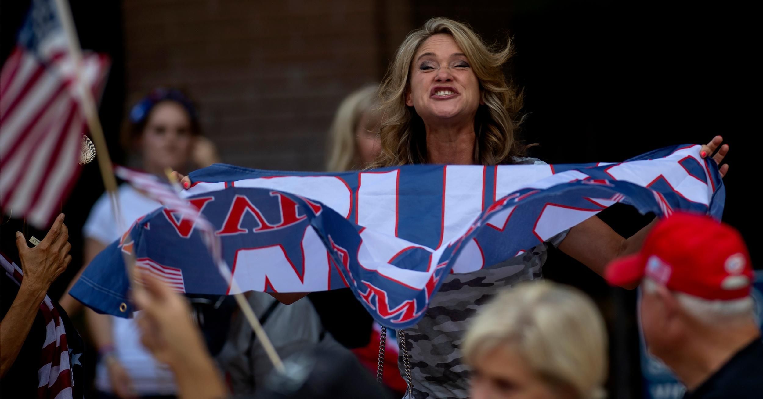 A woman holding a Trump flag reacts to a male driver in a passing car waving a Biden-Harris flag at an America First" rally.