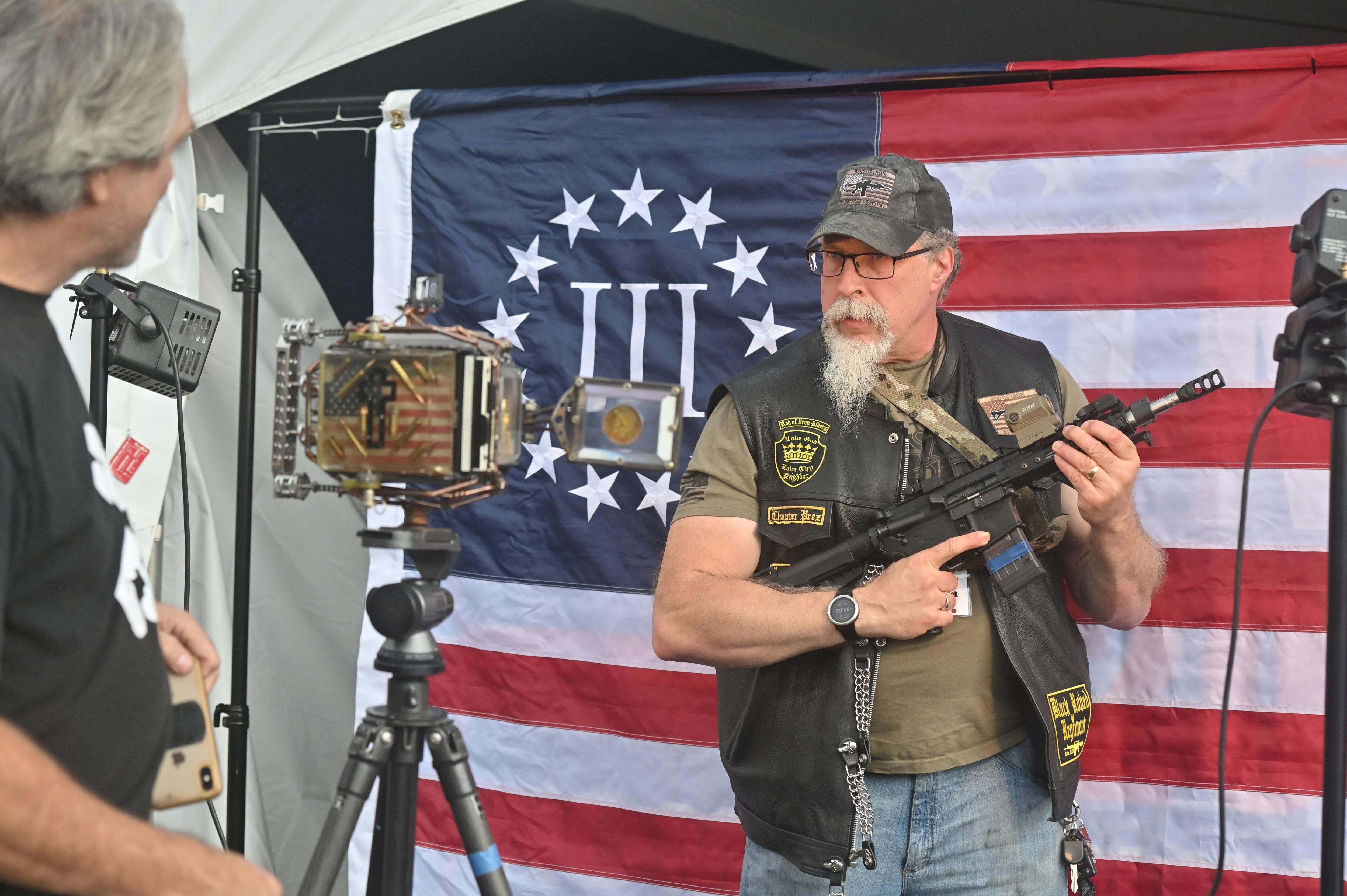A man holding an automatic rifle poses in front of a 3% flag, representing a far-right Christian fundamentalist worldview