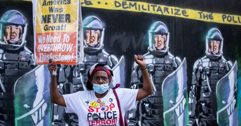 A protester wearing a mask holds a sign that reads, "American was never great! We need to Overthrow this system!" in front of a mural with New York Police Department officers in full riot gear and the banner above them that says, "Demilitarize The Police" during a march in Manhattan on July 18, 2020. (Photo: Ira L. Black/Corbis via Getty Images)