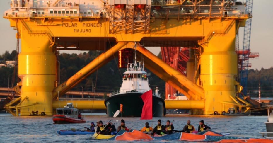 With the 40,000 ton Arctic drilling rig looming behind them, "kayaktivists" unfurl a large floating banner. (Photo: Greenpeace USA/ Flickr)