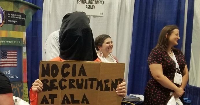 A protester holds a sign in front of the CIA's exhibitor booth at the American Library Association's 2019 annual conference. (Photo: Callan Bignoli)