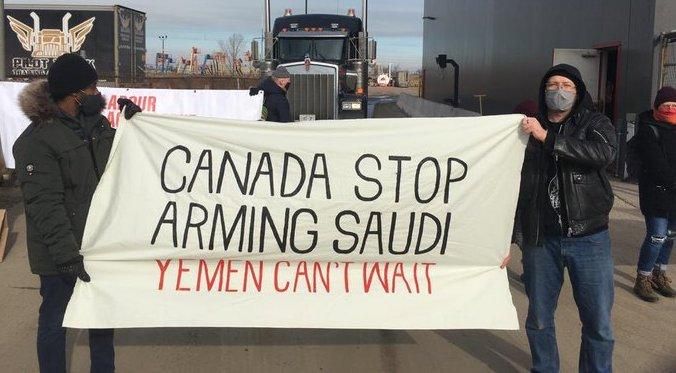People hold a banner and stand in front of trucks at Paddock Transport International in Hamilton, Ontario to stop the company from shipping what activists say are Canadian-made tanks to Saudi Arabia, on January 25, 2021.