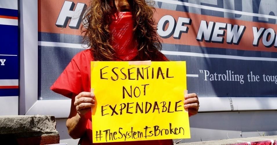 Person holds sign reading "Essential Not Expendable. #TheSystemIsBroken"