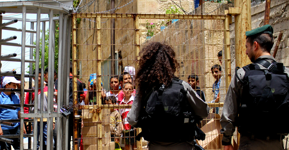  Two Israeli Soldiers guard a checkpoint in Hebron dividing zones H1 from H2 with Palestinian children standing at the other side of the fence. (Photo: June 2016, via Shutterstock) 